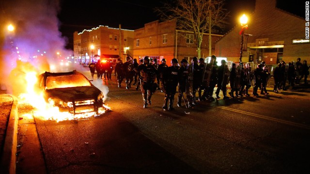 Police in riot gear move past a vehicle that continues to burn in Ferguson.