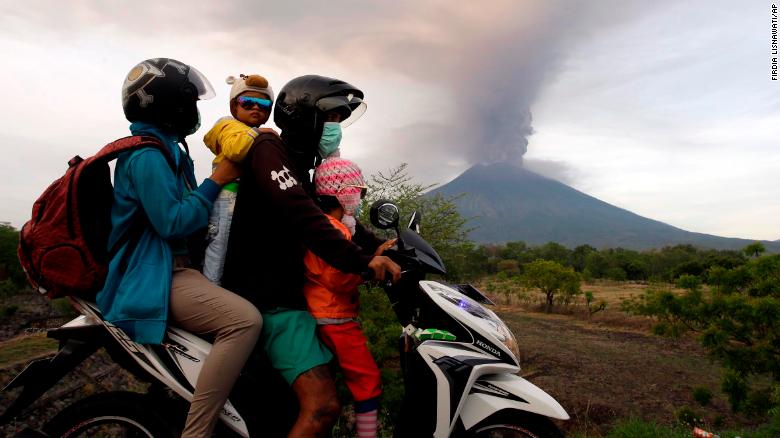 A family on a motorcycle passes by the Mount Agung volcano erupting in the background.