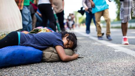 A girl sleeps on the side of the road as fellow migrants walk past.