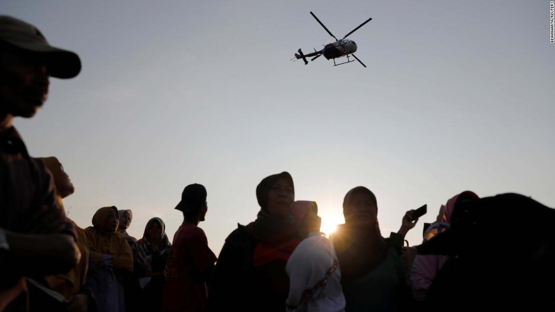 People watch a rescue team as a helicopter flies overhead.