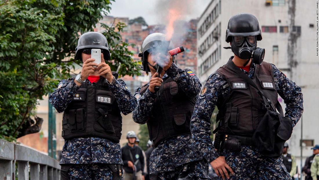 A National Police officer fires tear gas at demonstrators in Caracas while another shoots the scene with a cell phone.