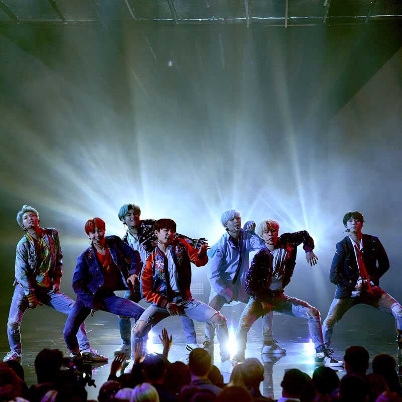 All seven members of BTS mid-dance move on a stage backlit by a white light wearing ripped jeans and jackets.