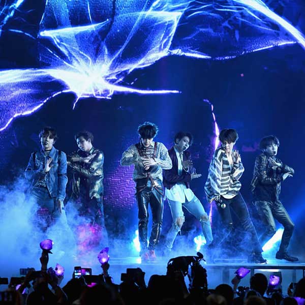 Six members of BTS performing on a stage bathed in blue light with electronic imagery as a backdrop.