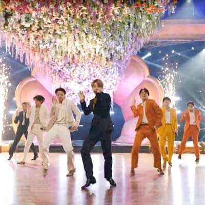 BTS members dance in colorful suits under a pastel backdrop on stage.