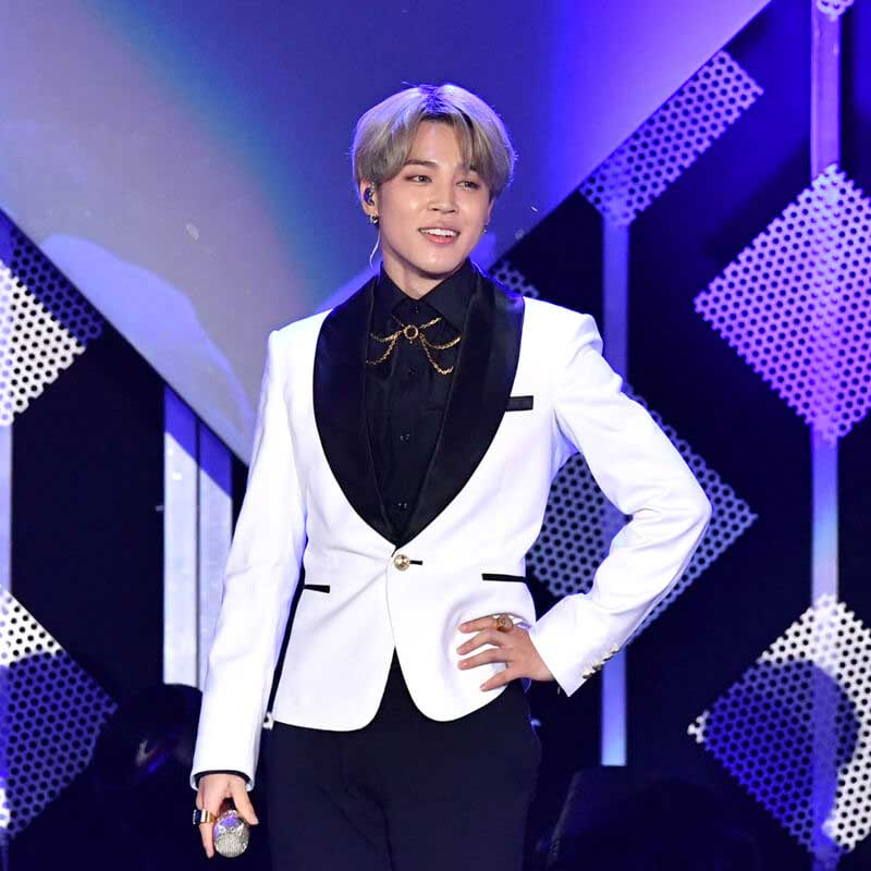 A member of BTS stands on stage holding a microphone in a white suit jacket with his hand on his hip.