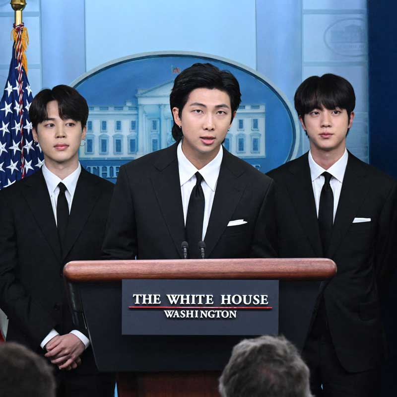 Three members of BTS in dark suits at a podium with the words ‘The White House Washington’ written on it.