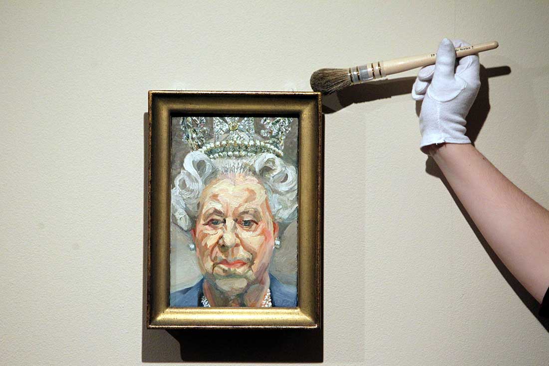 A gloved hand with a brush dusts a framed painting of the Queen.