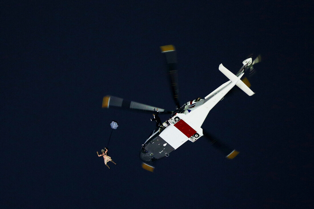 A view looking into a night sky with a helicopter and a a distant figure with a pink dress, jumping out in a spread-eagle position.