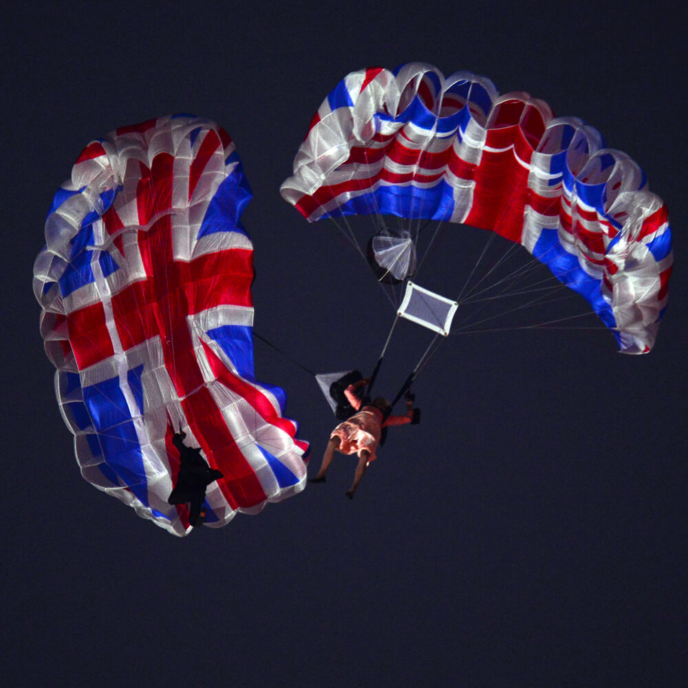 A view looking up at actors dressed as the Queen in a pink dress and James Bond in a suit parachuting. The parachutes are emblazoned with union jacks.