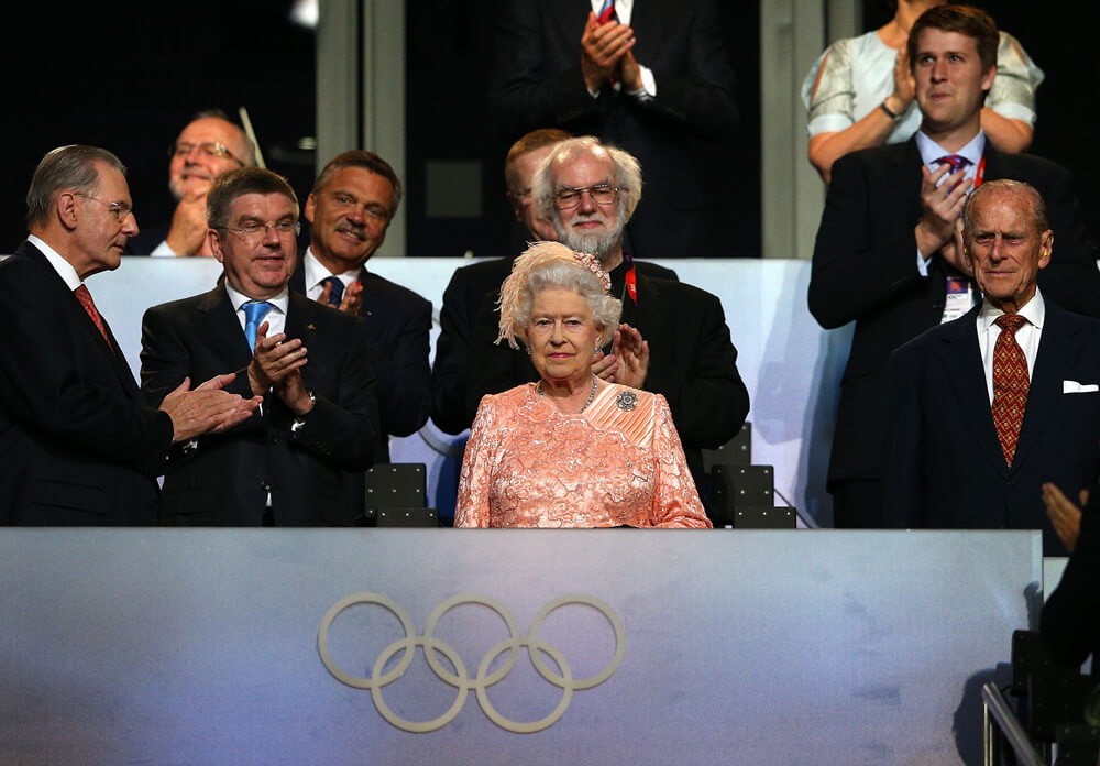 The Queen wearing a pink dress is sitting behind the Olympic rings, surrounded by dignitaries applauding.