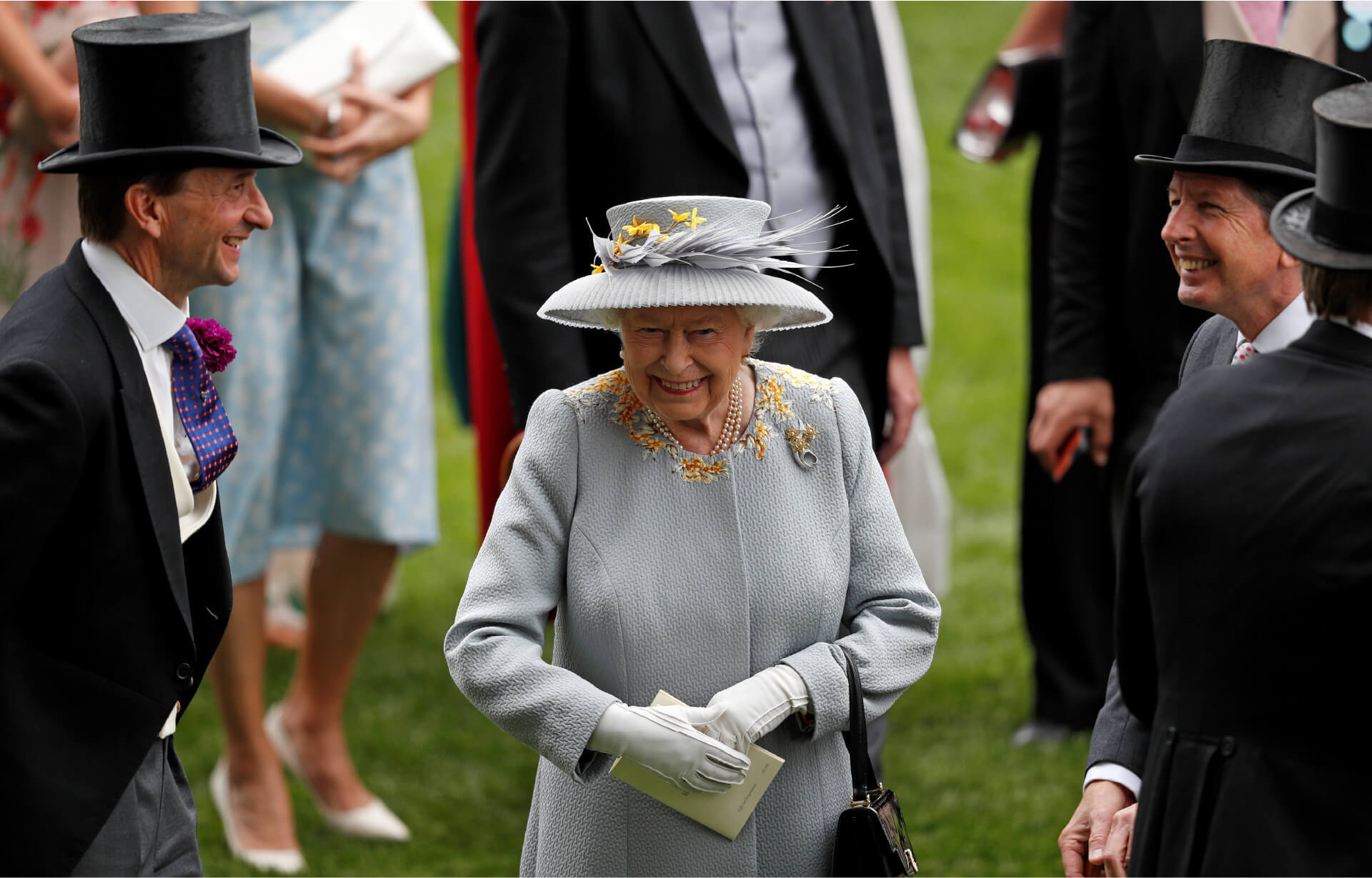 Queen Elizabeth wearing a grey coat embroidered with yellow flowers around the collar, with a matching medium brimmed grey hat that is adorned with feathers and yellow flowers. She is surrounded by men in black top hats and tails.