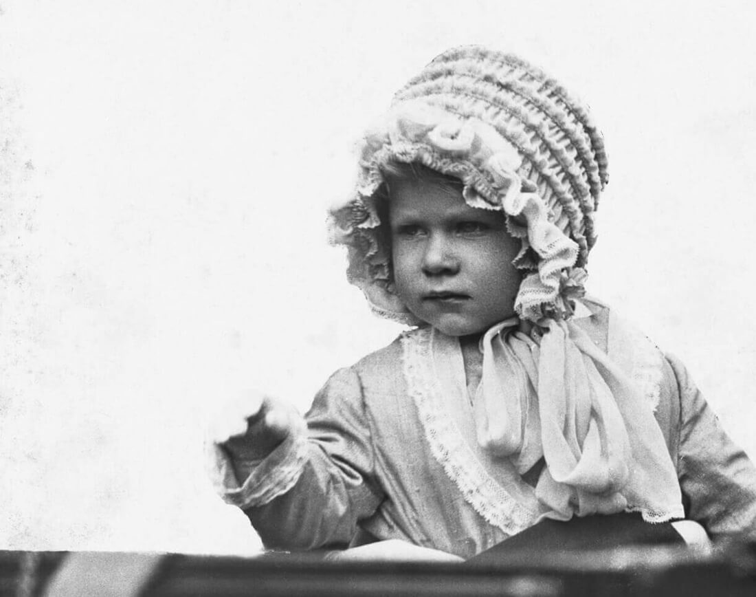 A baby Elizabeth sitting up in a carriage wearing an elaborate lace bonnet tied with a bow beneath her chin and a little coat trimmed with lace.