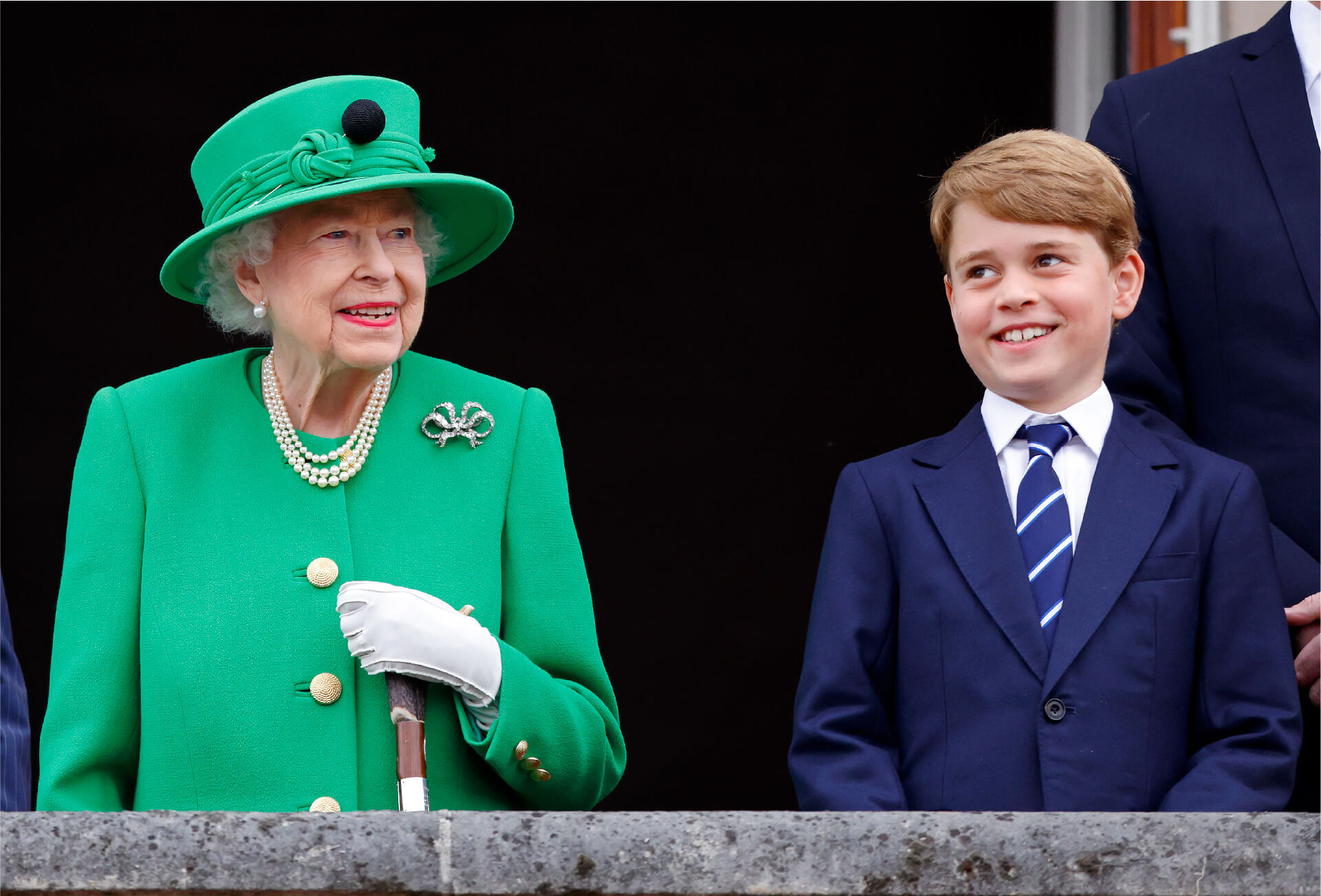 Queen Elizabeth standing on a balcony next to a smiling young Prince George.  The Queen wears an emerald green suit jacket with a matching wide-brimmed hat, a three-row pearl necklace and a diamond brooch.  She leans on a cane which she grasps with a white-gloved hand.
