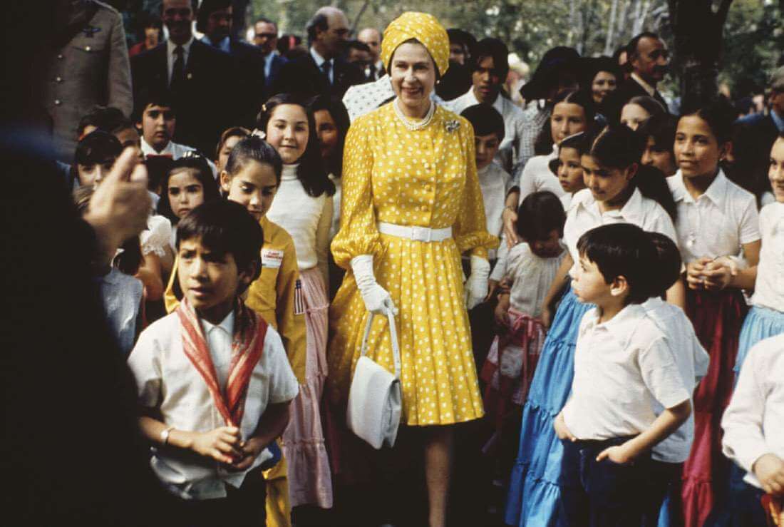 महारानी एलिजाबेथ स्कूली बच्चों की भीड़ के बीच एक चमकीले पीले रंग की पगड़ी-शैली की टोपी पहने हुए हैं, जिसमें एक सफेद बेल्ट के साथ कमर पर सिनी हुई पोल्का-बिंदीदार पोशाक है।