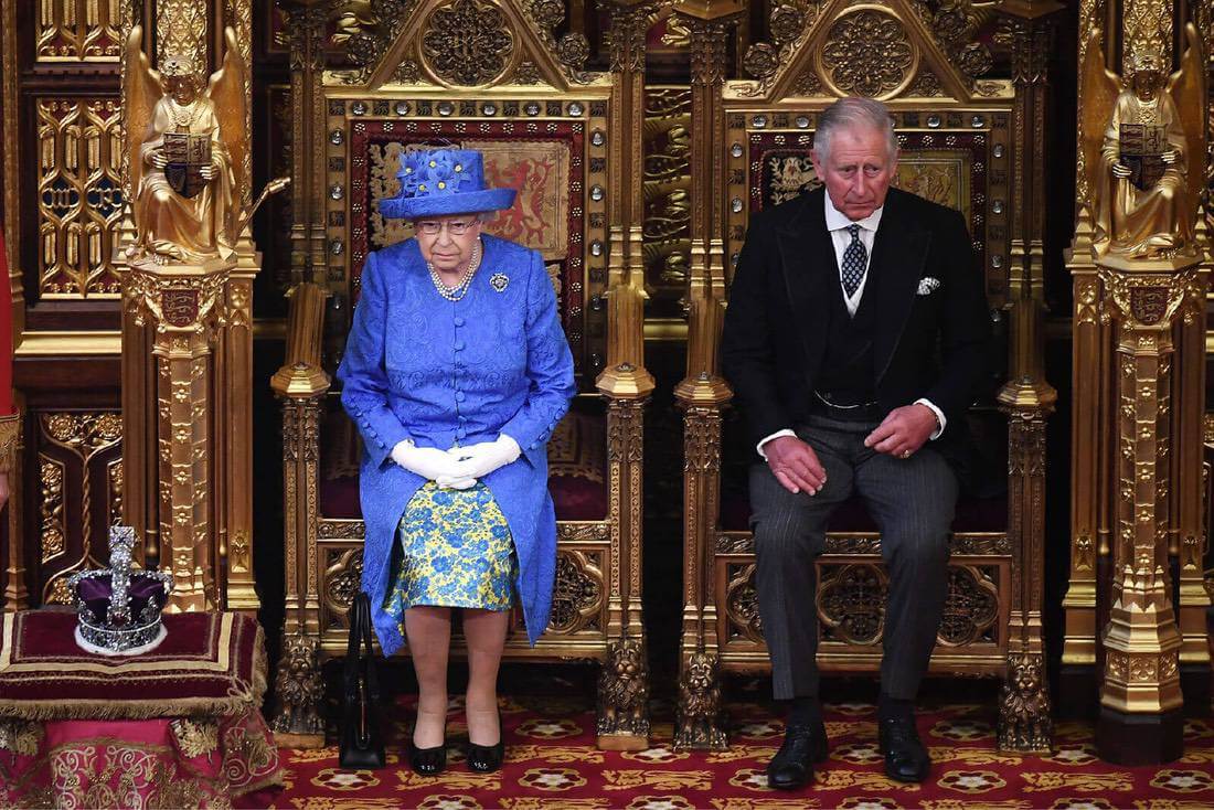 Queen Elizabeth sits on a golden throne in Parliament next to her son Prince Charles who wears a morning suit. The Queen wears a royal blue hat with yellow flower details and a matching buttoned coat and dress.