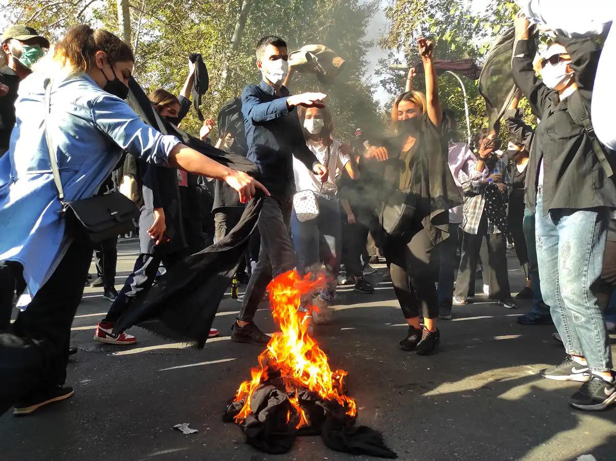 A group of young people wearing medical masks jump with their fists in the air around a pile of black material that has been set on fire.