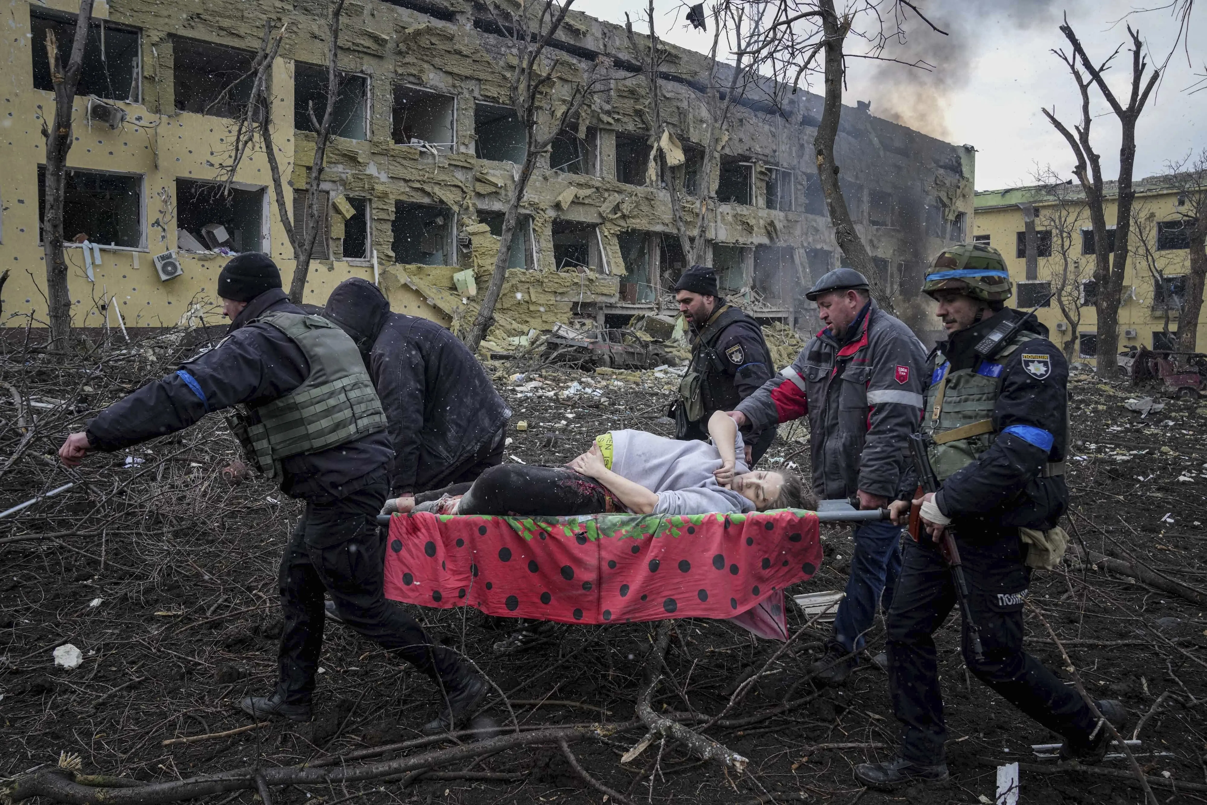 A pregnant woman is carried on a stretcher by five men amid smoking rubble and a bombed out two-story building in the background with charred trees.