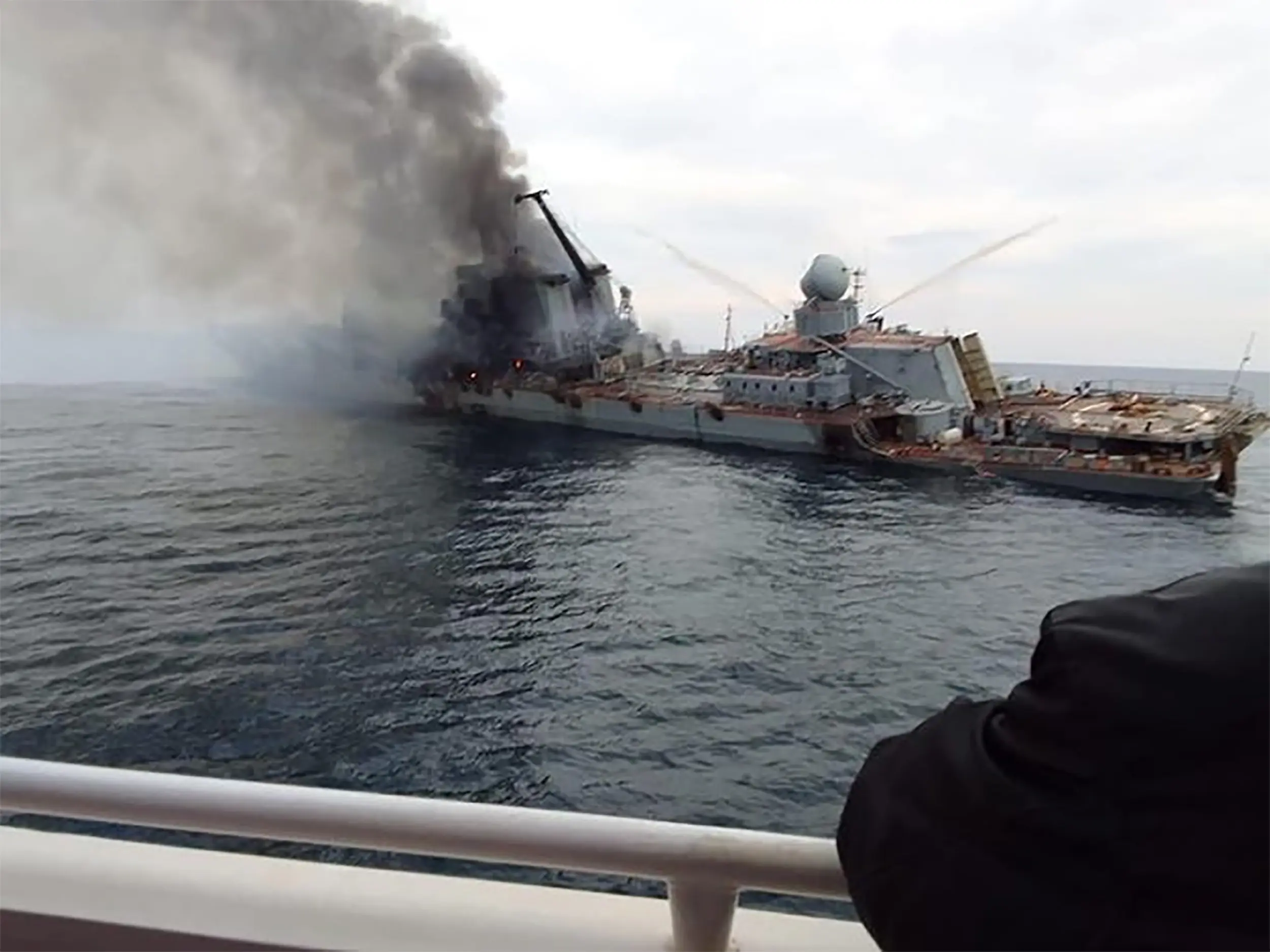 A huge military ship on the water is on fire with dark grey smoke coming out of it.