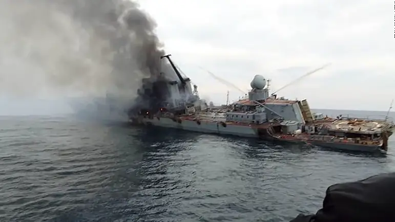 A huge military ship on the water is on fire with dark grey smoke coming out of it.