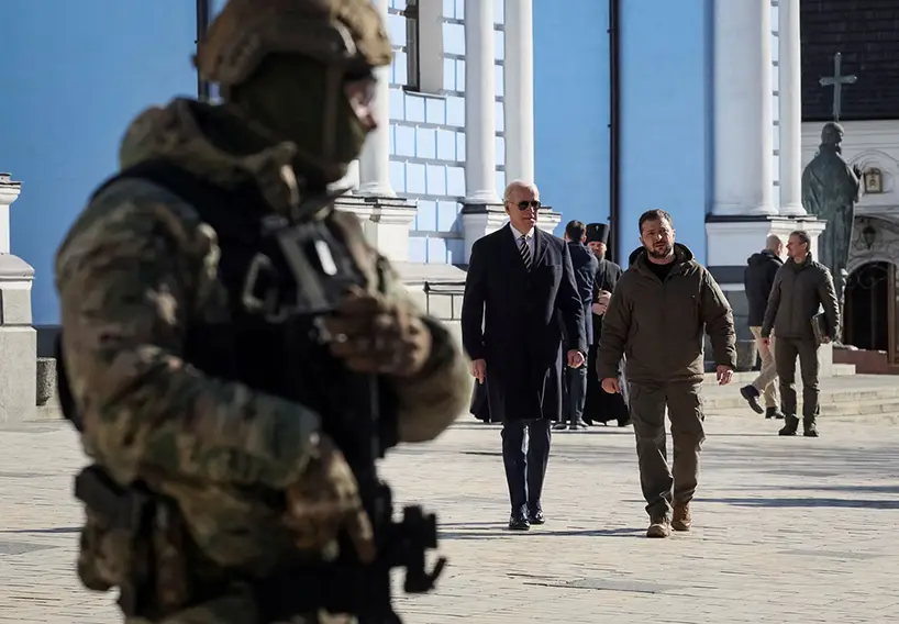 President Biden wearing a coat and sunglasses and President Zelensky wearing his signature dark green pants and jacket walk across a square with an armed soldier in front of them.