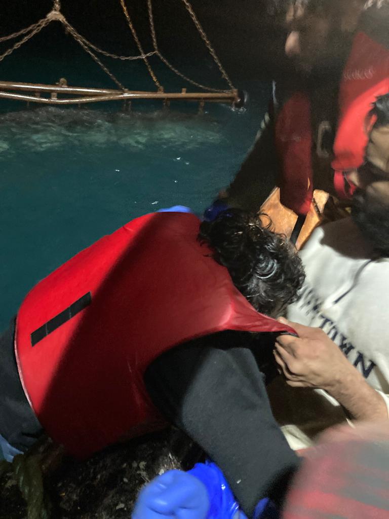 A man is pulled by the top of his lifejacket into a boat by another man. The sea is in the background and the sky is pitch black.