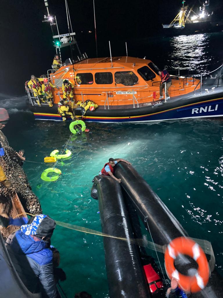 An orange lifeboat with at least six rescuers haul a man in from the sea water. A black rubber dinghy a few metres away from the lifeboat has a man wearing a lifejacket clinging to it.