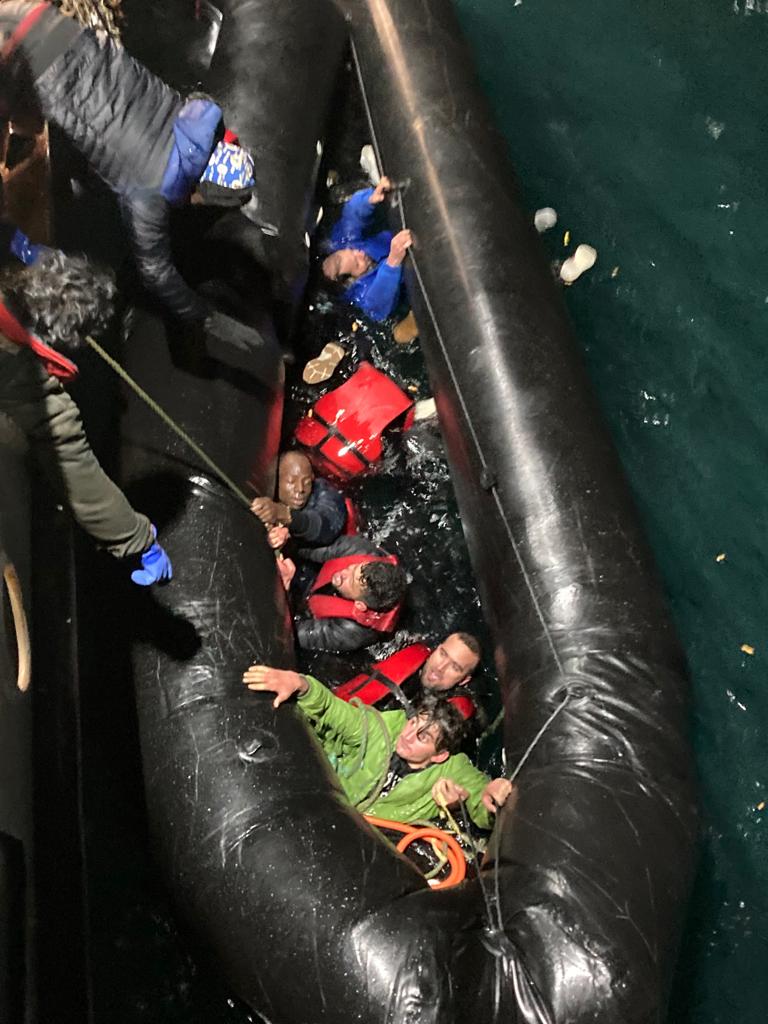 Many young men, some in lifejackets, crammed into a small black rubber boat in the sea looking distressed as they reach up to rescuers to help them from another boat.