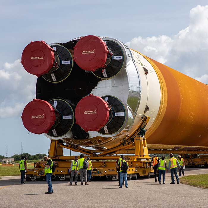 A huge rocket on its side being transported on a road dwarfs people in the foreground.