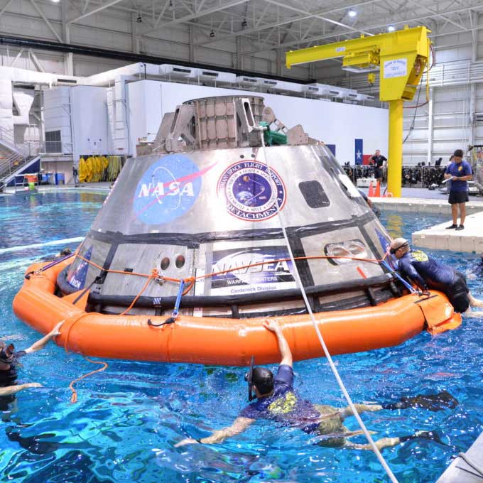 Silver Orion spacecraft floats on an orange inflatable in a pool with people swimming alongside.