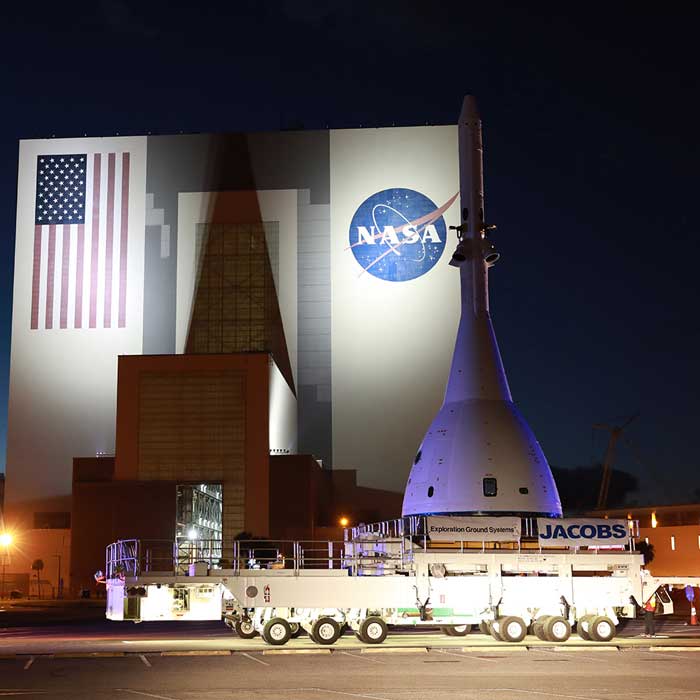 A test version of Orion carried by a transporter, in front of the Vehicle Assembly Building.