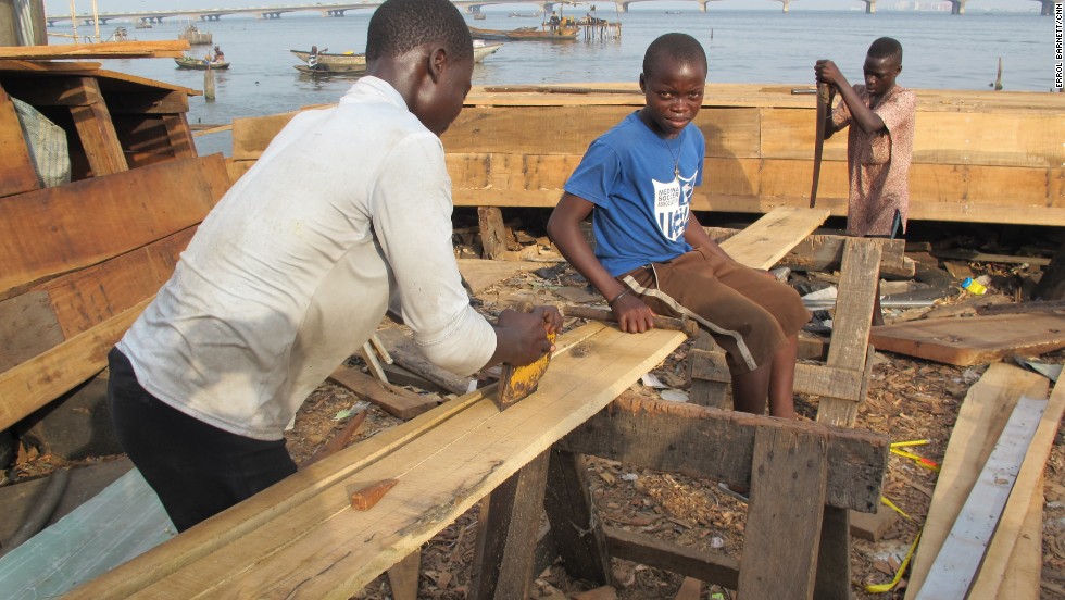 Young men in Makoko are typically put to work building canoes, in business ventures supervised by elders. Here, one teen sits on a plank of timber as two others work to saw it into pieces.