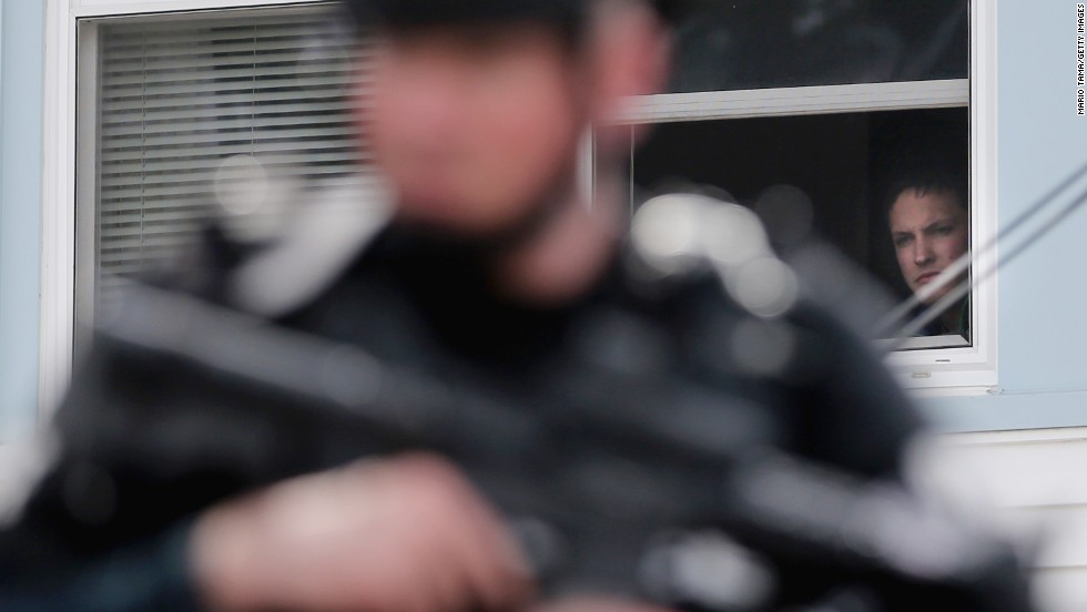 A man watches from the window of a home as a SWAT team member keeps watch on Friday, in Watertown, Massachusetts.