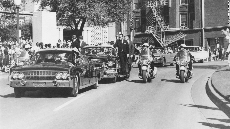 Seen through the limousine&#39;s windshield as it proceeds along Elm Street past the Texas School Book Depository, Kennedy appears to raise his hand toward his head after being shot. The first lady holds Kennedy&#39;s forearm in an effort to aid him. 