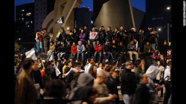People on top of the base of a monument watch and cheer as protesters walk during a march in Mexico City.