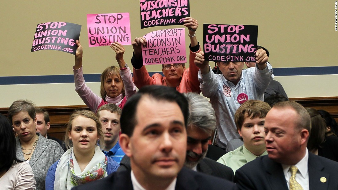 Members of Code Pink (left to right) Medea Benjamin, Liz Hourican and Tighe Barry, hold signs to protest as Walker (center) takes his seat during a hearing before the House Oversight and Government Reform Committee April 14, 2011, on Capitol Hill in Washington, D.C.