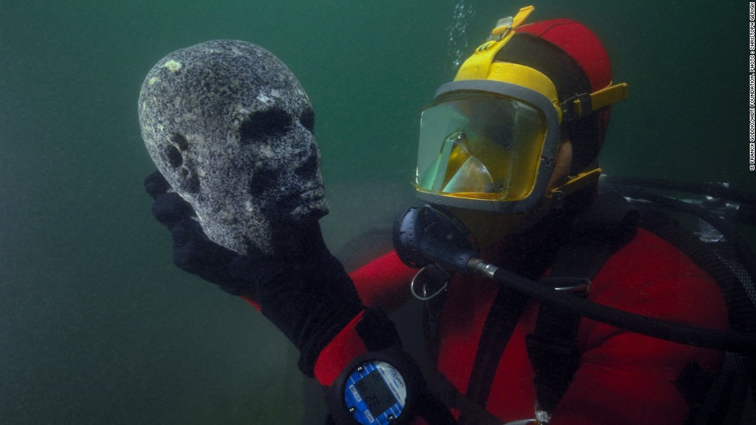 Here, a diver inspects a recovered black granite statue depicting a priest&#39;s head. The scalp is probably shaven, as was befitting of priests, for reasons of purity. The cheekbones are pronounced and the cheeks hollowed. On the forehead, the scarification mark of priests is visible and the eyes, now hollowed out, would have been incrusted with something unknown today. 