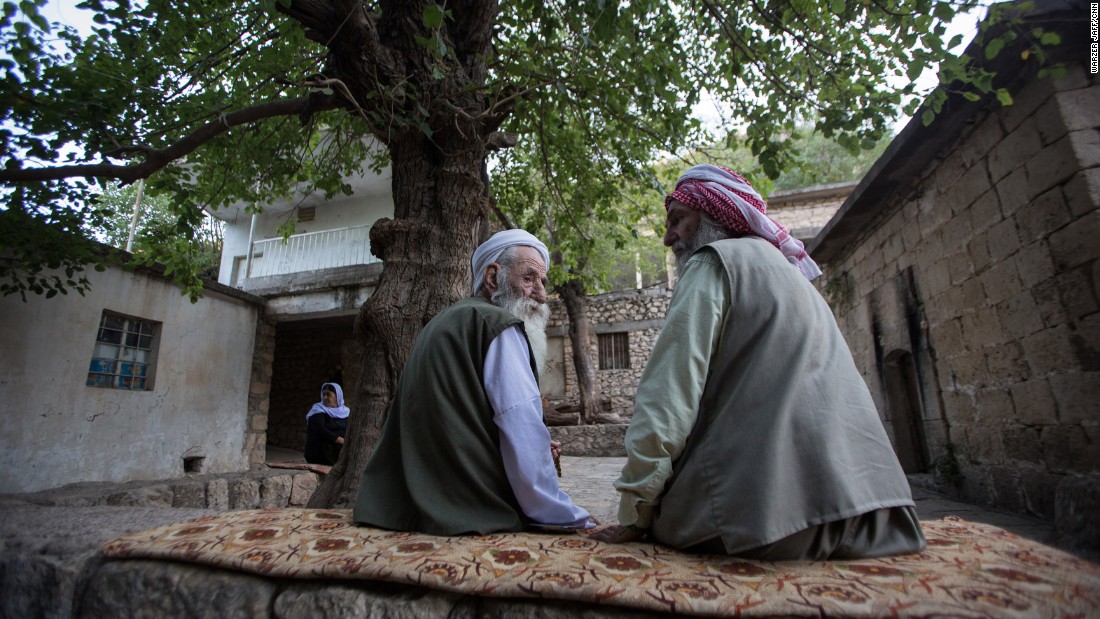 Two older Yazidi men talk under a tree.