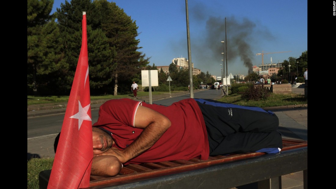 A protester rests on a bench as smoke billows from the Turkish military headquarters in Ankara.  