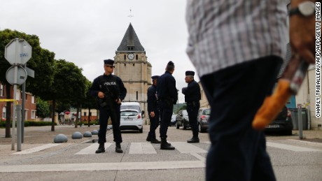 A man carries a French baguette as police officers stand guard the Saint-Etienne church.