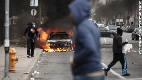 People walk past burning cars near the intersection of Pennsylvania Avenue and North Avenue in Baltimore on April 27, 2015. Riots erupted in the city following the funeral service for Freddie Gray, who died while in Baltimore Police custody.