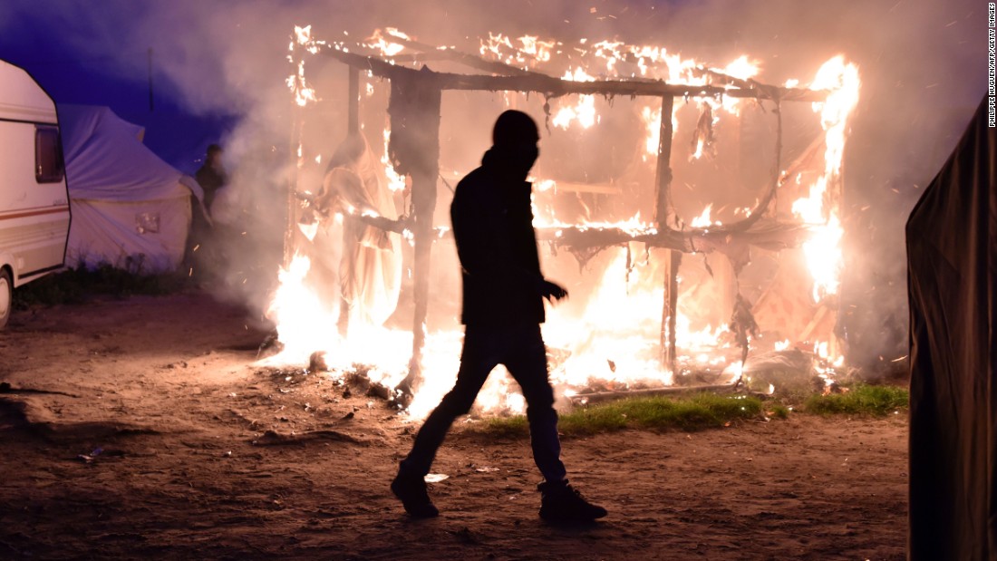 A migrant walks past a burning shack that was set on fire, as a demolition crew began tearing structures down on Tuesday, October 25.