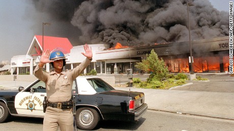 A California Highway patrolman directs raffic around a shopping center engulfed in flames in Los Angeles, 30 April 1992. Riots broke out in Los Angeles, 29 April 1992, after a jury acquitted four police officers accused of beating a black youth, Rodney King, in 1991, hours after the verdict was announced.        (Photo credit should read CARLOS SCHIEBECK/AFP/Getty Images)