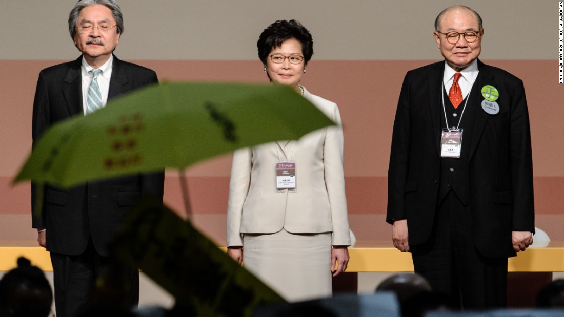 Carrie Lam (center) stands on stage after being selected to be the next Hong Kong Chief Executive on March 26, 2017. In the foreground, a yellow umbrella is unfurled to protest the undemocratic nature of the race Lam won. 