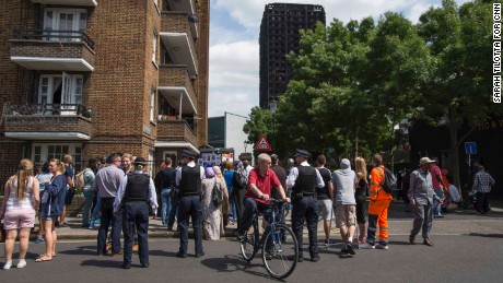 A crowd gathers in view of the blackened Grenfell Tower.