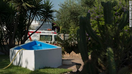 An ambulance is seen through a neighbor&#39;s front lawn, where a hose dangles in a half-empty pool.