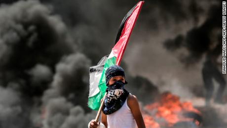 TOPSHOT - A Palestinian boy holding his national flag looks at clashes with Israeli security forces near the border between the Gaza Strip and Israel east of Gaza City on May 14, 2018, as Palestinians protest over the inauguration of the US embassy following its controversial move to Jerusalem. - Dozens of Palestinians were killed by Israeli fire on May 14 as tens of thousands protested and clashes erupted along the Gaza border against the US transfer of its embassy to Jerusalem, after months of global outcry, Palestinian anger and exuberant praise from Israelis over President Donald Trump&#39;s decision tossing aside decades of precedent. (Photo by MAHMUD HAMS / AFP)        (Photo credit should read MAHMUD HAMS/AFP/Getty Images)