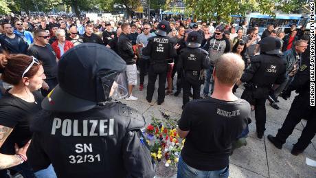 Riot police and citizens stand next to a makeshift memorial marking the site where three people were injured in a fight early Sunday. One man later died from his injuries, police said.