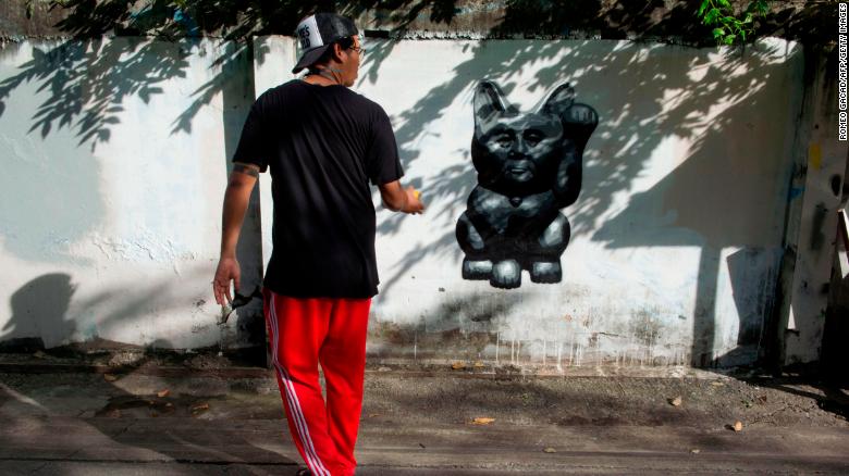The Thai street artist who goes by the name of &quot;Headache Stencil&quot; walks next to his graffiti caricature of Thailand&#39;s junta chief depicted as a &quot;lucky cat&quot; with paw raised to rake in money, spray-painted on a fence in Bangkok. 