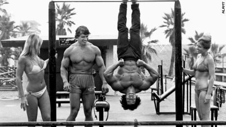 Arnold Schwarzenegger stands next to Franco Columbu, hanging upside down, in this image from 1977.
