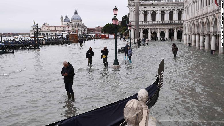 People walk past a stranded gondola across the flooded Riva degli Schiavoni embankment and St. Mark&#39;s Square in the background.